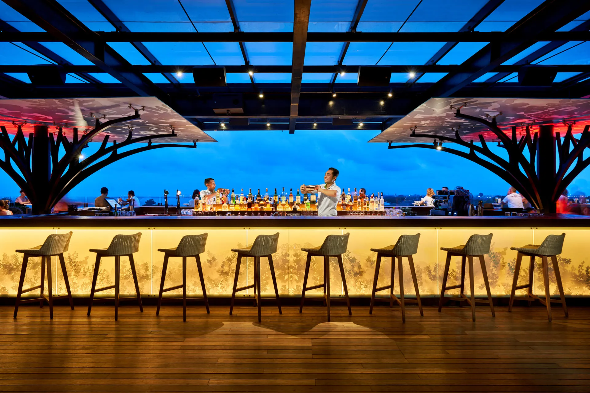Bar at blue hour with bartender, woven stools and tropical foliage backlit panel — Above Eleven Bali bar photography